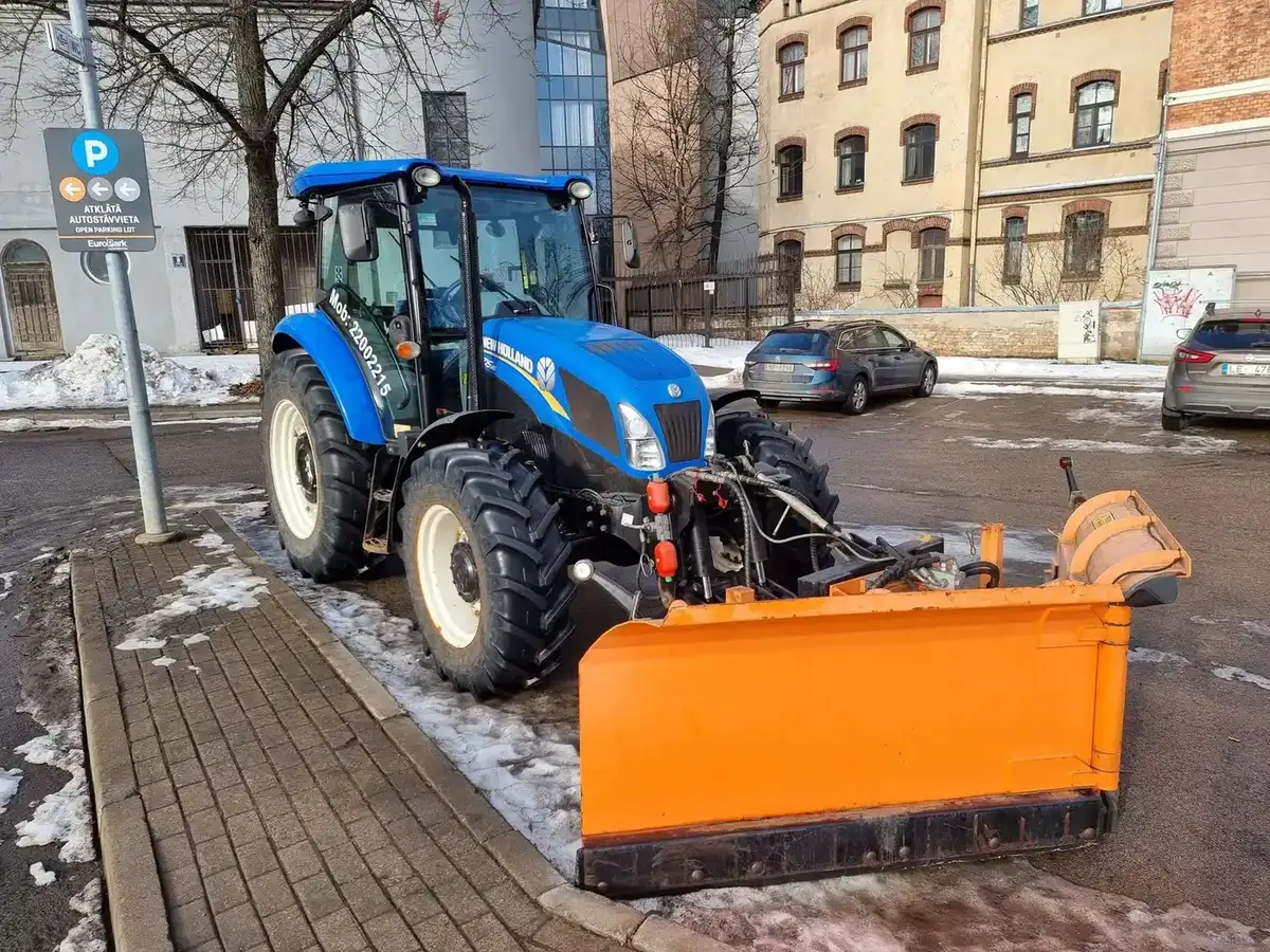 Трактор с оранжевым снегоуборочным отвалом чистит парковку EuroPark в центре Риги